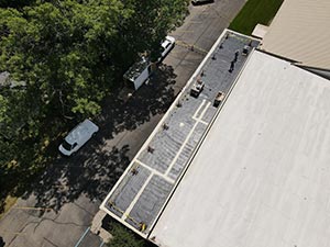 Commercial-Roofing-Services-Indianapolis-IN-Indiana-1 | CVC Roofing & Sheet Metal Aerial view of a flat roof with solar panels along the edge of a building, with a tree line and a street below including a parked white car.